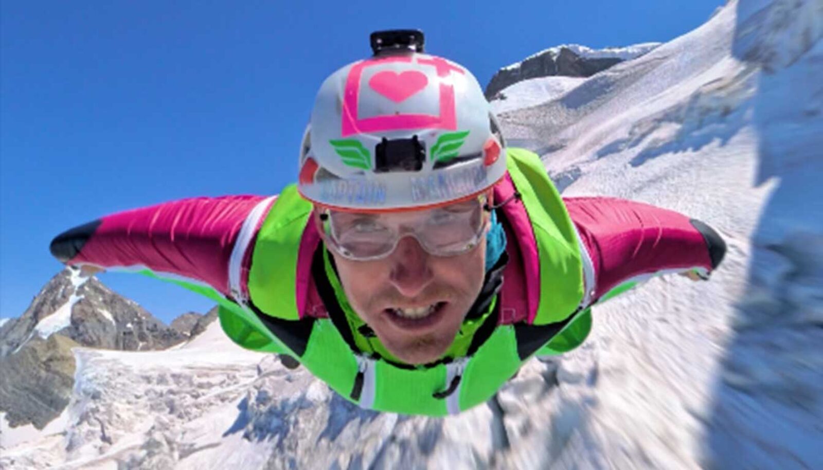 Wingsuit pilot in mid-flight, direct to camera, over an alpine landscape