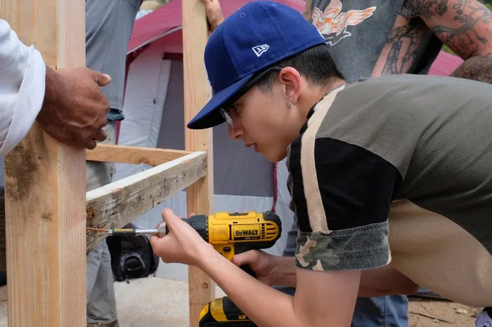 A youth in safety goggles uses a DeWalt drill to build camp structures at Freedom Land - Camp Care More life skills