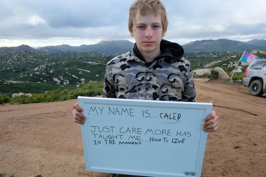 Caleb holds a whiteboard sign at Freedom Land that reads: Just Care More has taught me how to live in the moment
